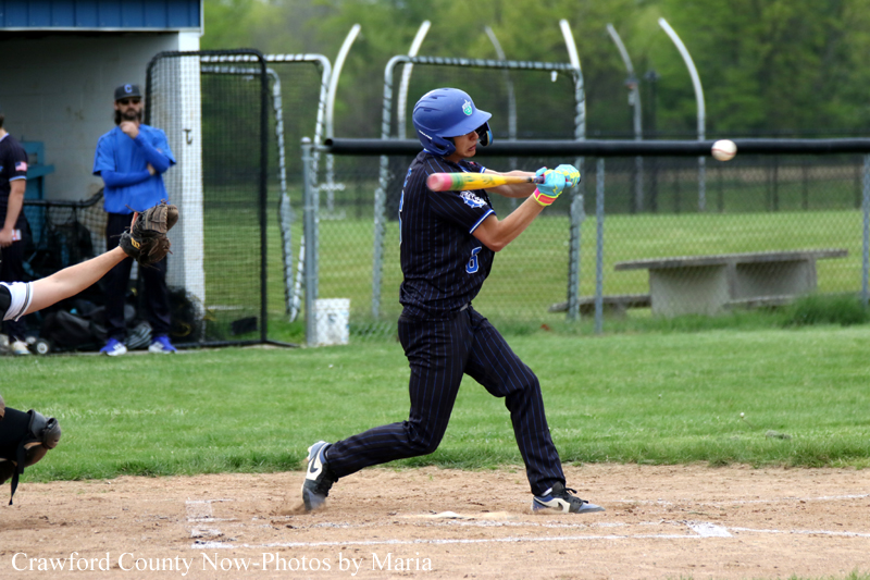 A baseball batter in navy uniform swings at a pitch during a game, with a catcher partially visible on the left and players/fence in the background