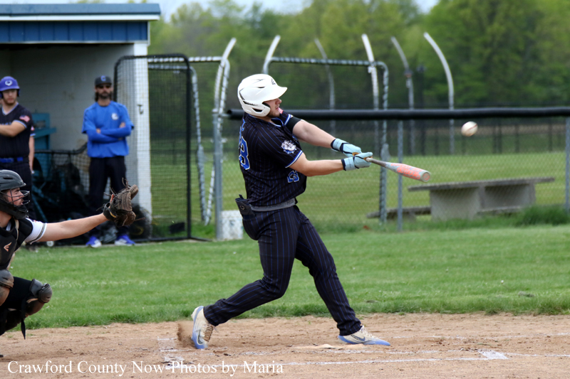 Baseball batter in navy uniform swings at a pitch during a game, glove on front foot ready to run.