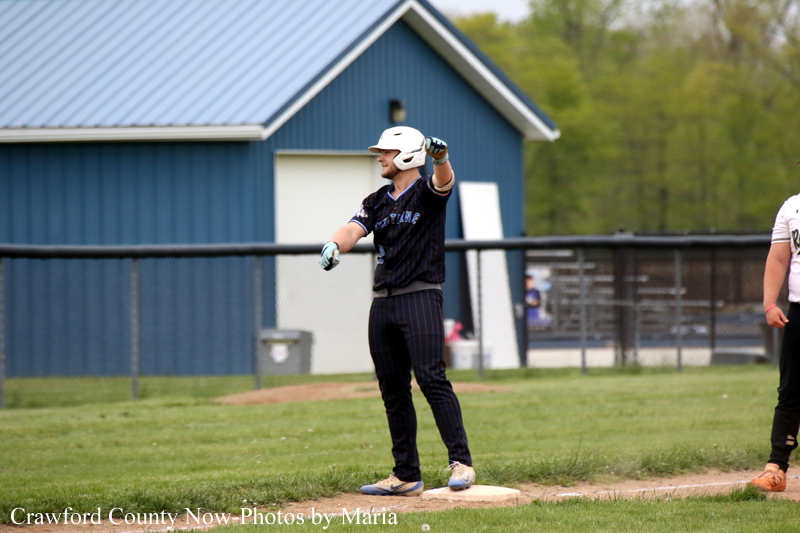 Baseball player in a dark uniform and white helmet stands on a base, pointing toward the infield during a game with a blue building in the background.