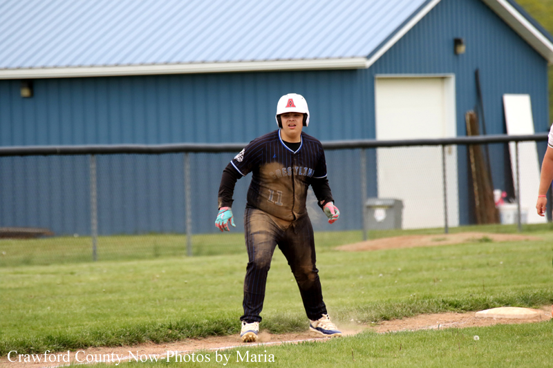 Young baseball player in a dark blue uniform stands on a base, helmet on, dirt-smeared uniform, ready on the field with a blue building in the background.