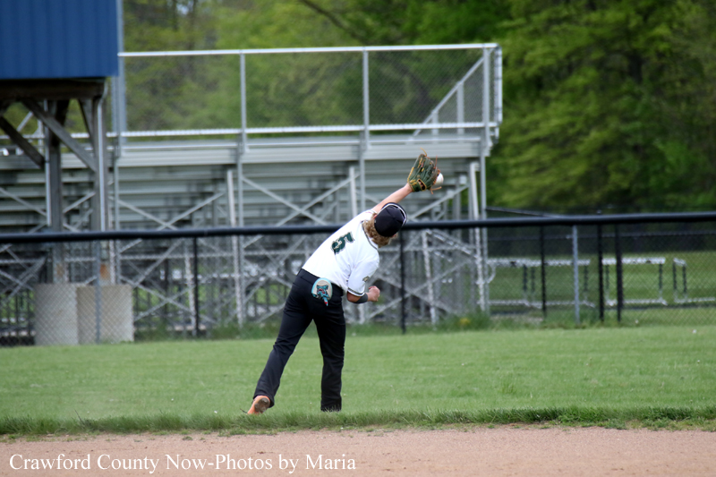 Baseball fielder in white uniform reaching with glove to catch a ball in the outfield, bleachers in the background.