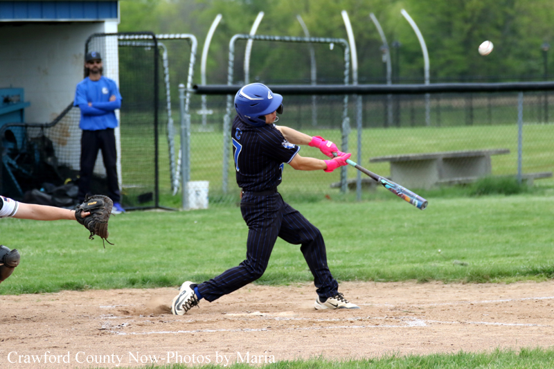 Baseball batter in a dark navy uniform with pink gloves swinging at a pitched ball on a dirt infield; defender's glove visible on the left and a chain-link fence in the background.
