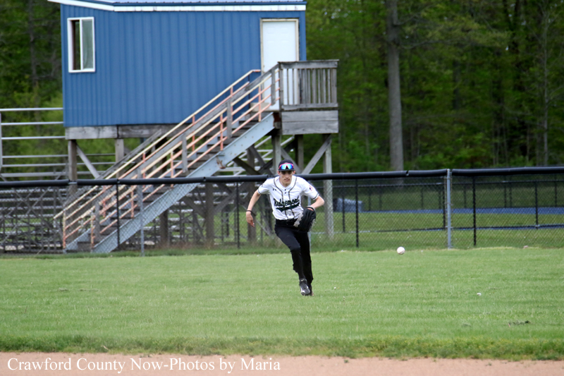 Baseball player in white jersey runs on the outfield grass with a blue elevated building and stairs behind a chain-link fence.