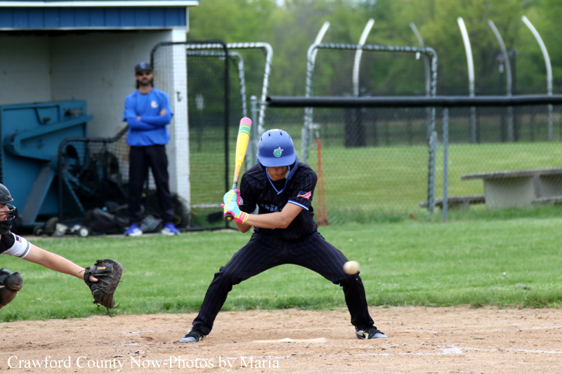 Baseball batter in dark uniform and blue helmet swings at a pitch at home plate, catcher glove ready nearby in the infield dirt.