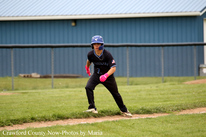 Baseball player in black uniform wearing pink gloves stands on a dirt basepath, ready to run, with a blue outfield building in the background.