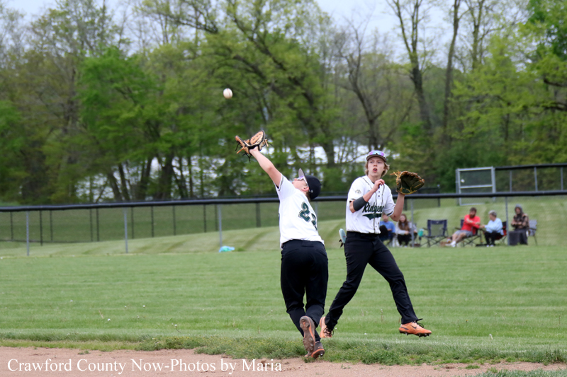 Two softball players in white jerseys jump to catch a ground ball on a green field, with a fielder glove reaching up and a teammate ready nearby.
