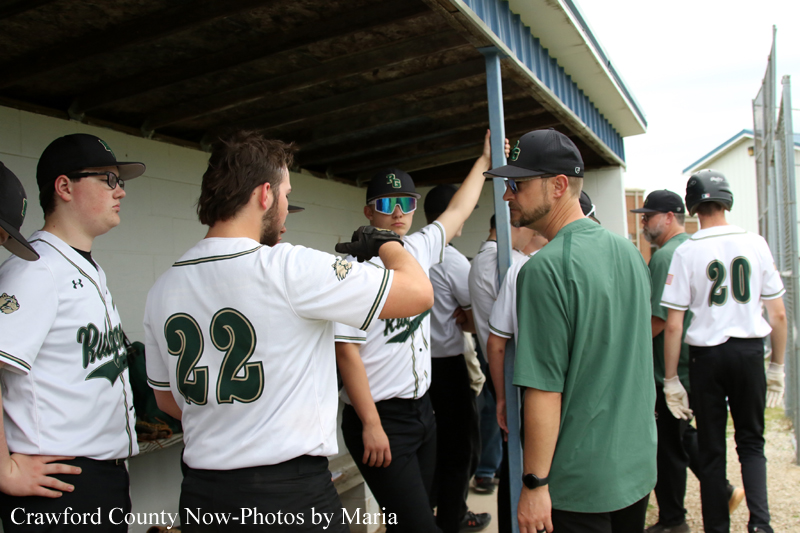 Baseball players in white and green uniforms gather in a dugout; a coach in a green shirt talks to them as teammates wait nearby.