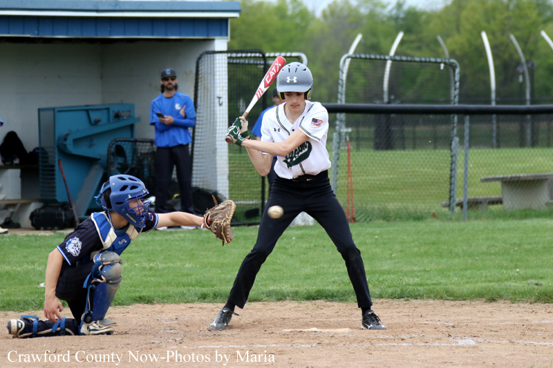 Young female softball batter at home plate swings as the ball approaches; catcher in blue gear crouches behind, with a coach and fence in the background.