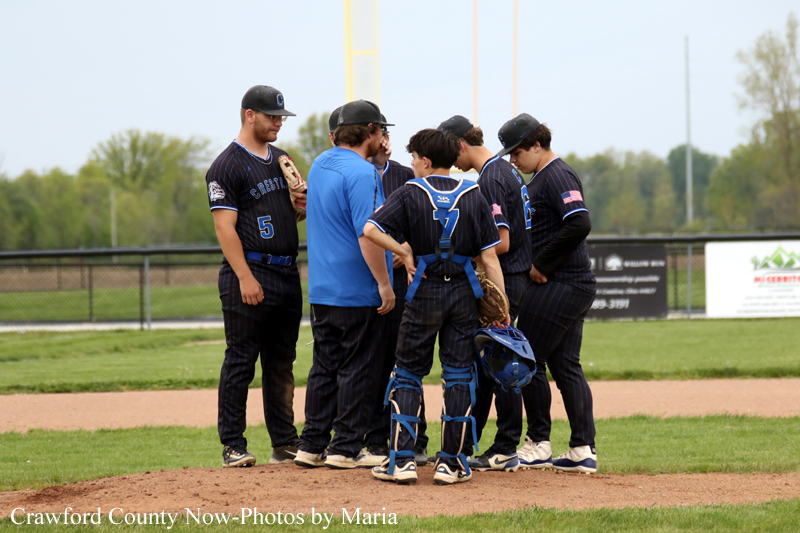 Group of baseball players and a coach in a huddle on the infield dirt during a game on a sunny day.