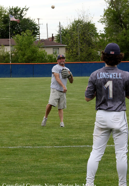 Baseball scene: a pitcher throws a ball toward a batter on a green field, with trees and a fence in the background.