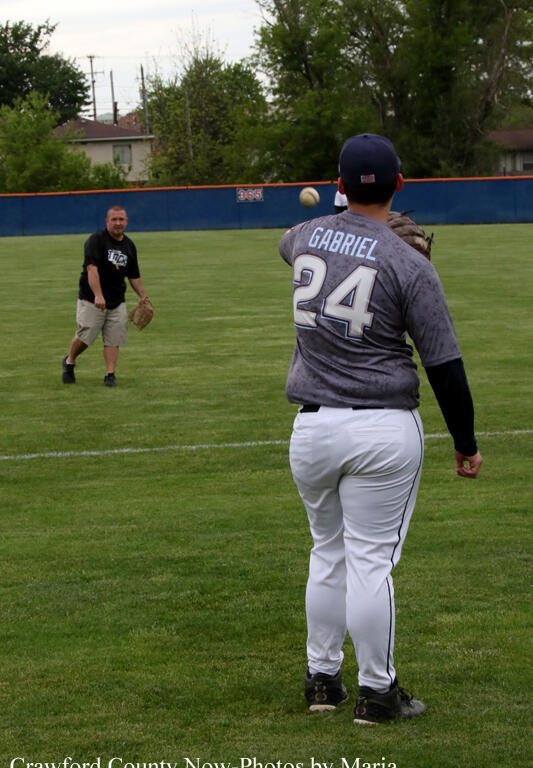 Baseball pitcher wearing number 24 with the name Gabriel on his gray jersey winds up to throw a pitch on a grassy field, with a fielder in black in the background.