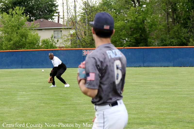 Baseball game scene: pitcher in gray uniform with number 6 faces away from the camera as another fielder moves to field a ball on the grass.