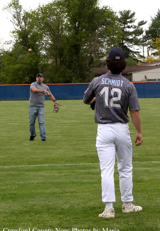 Baseball field with two players: foreground player in a gray Schmidt #12 jersey facing away; in the distance a teammate reaches to catch a throw on the grass.