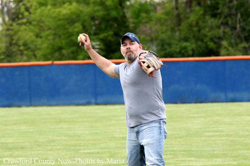 Adult man in a gray t-shirt and cap throws a baseball on a grassy field, with a blue outfield fence in the background.