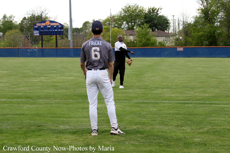 Two baseball players on a grass field with a blue outfield wall; foreground player faces away, gray jersey with number 6, white pants, and cap. A teammate stands in the infield with a glove, gesturing as if guiding plays.