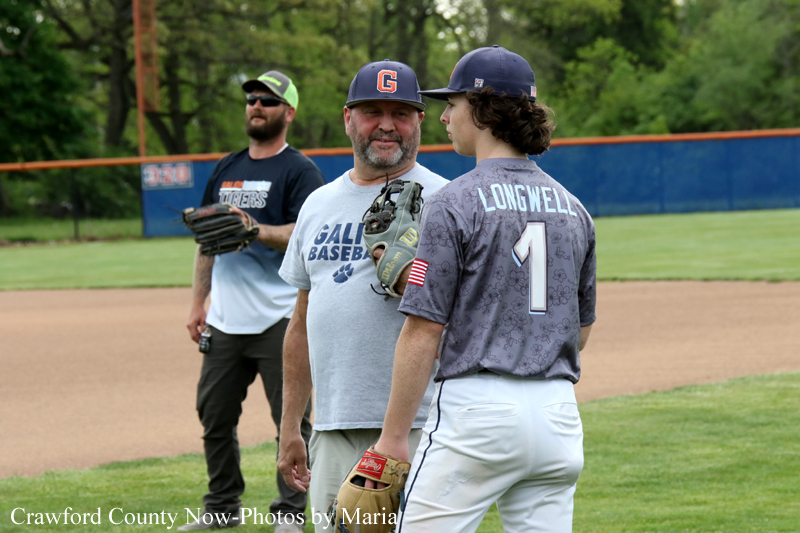 Three men on a baseball field, two players chatting near the infield with gloves on, one wearing a cap with a G logo and a gray 'GAL... BASEBALL' shirt nearby, daytime scene