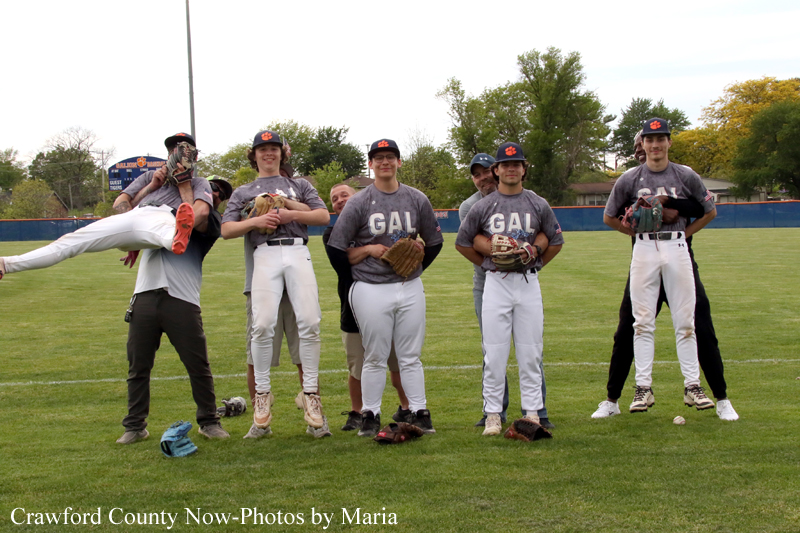 Seven baseball players pose for a team photo on a grassy field, wearing gray GAL shirts and white pants with gloves on.