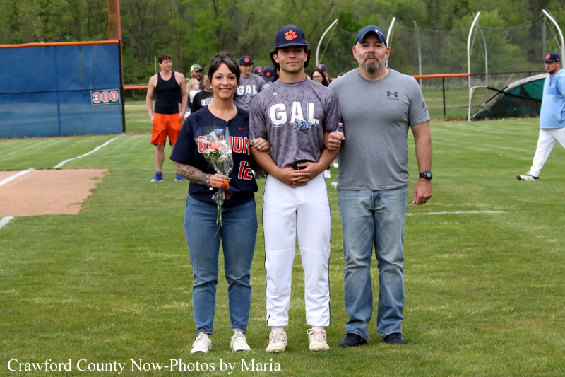 Family poses with a young baseball player in a gray GAL jersey on a baseball field after a game; woman holds flowers, man standing with him, others in background.