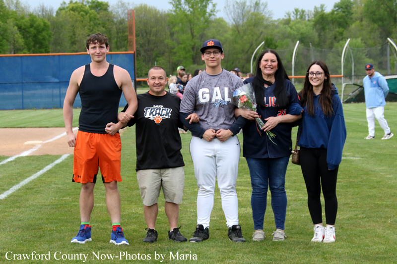 Six people stand together on a baseball field, smiling for a group photo; some wear team uniforms, one holds flowers.