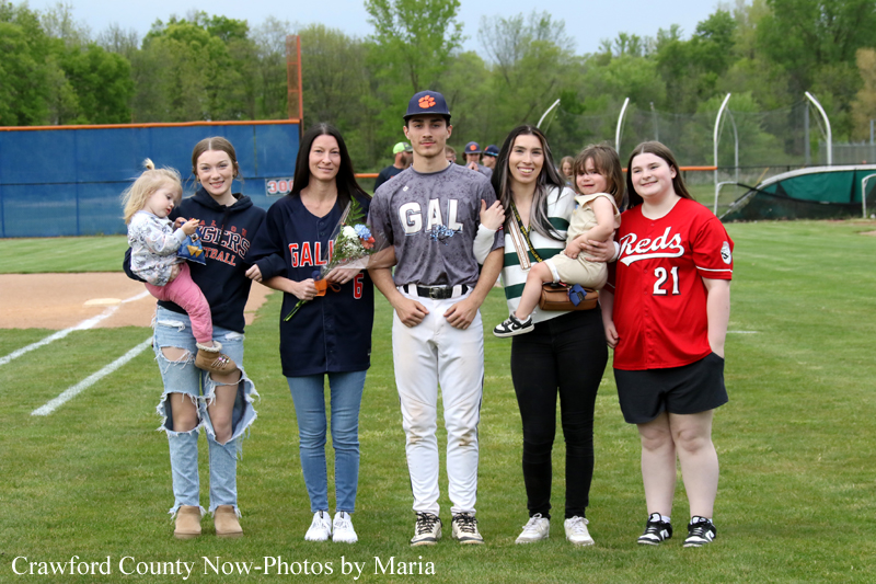 Group of teammates and family posing on a baseball field, several wearing GAL uniforms and one in a Reds jersey, with children in arms in the center and on the ends.