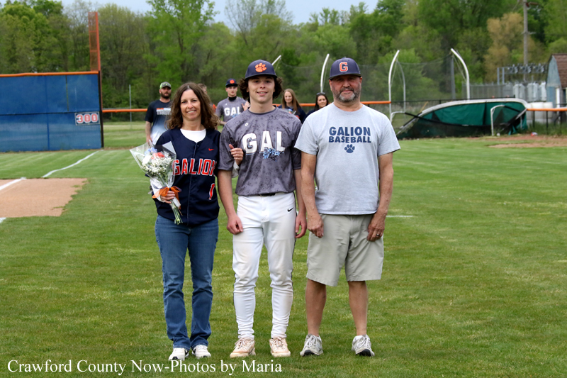 Three people stand on a baseball field: a player in Galion uniform between a woman with a bouquet and a man in a Galion Baseball shirt, posing for a photo.