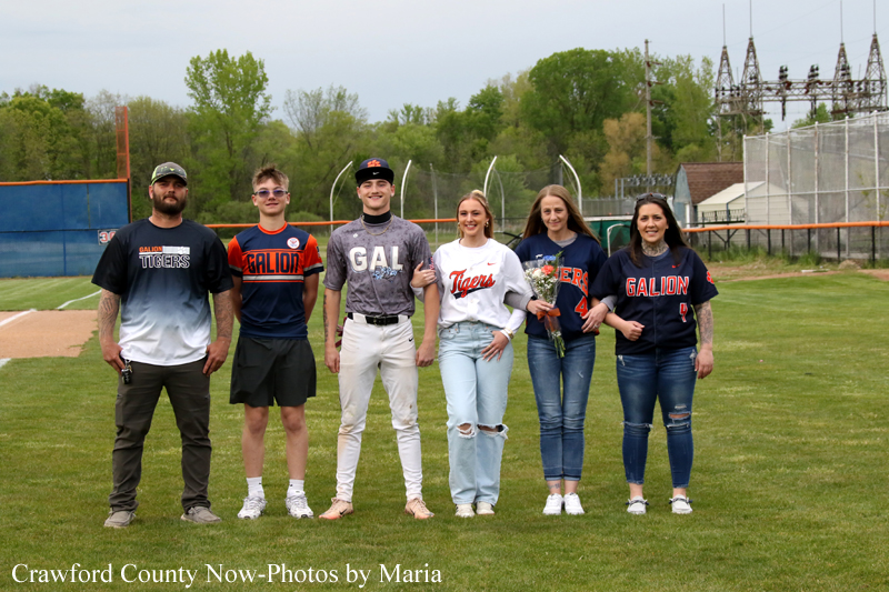 Group of six young people in baseball uniforms posing for a photo on a grassy field, with jerseys showing Galion and Tigers logos, in front of a backstop and fence.