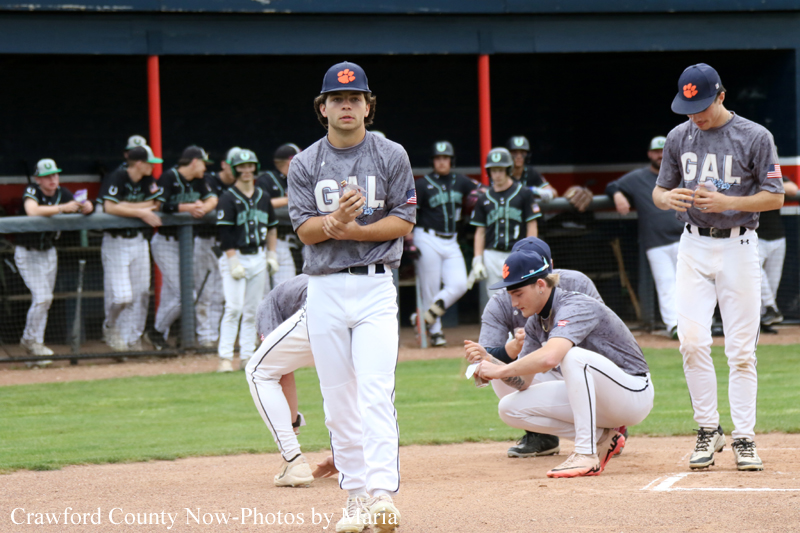 Baseball scene with a pitcher in gray uniform standing on the mound, glove ready, while teammates watch from the dugout in the background.