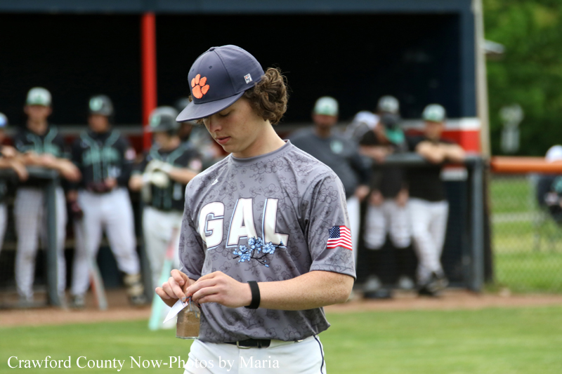 Baseball player in gray GAL jersey adjusts a small bottle in a field with teammates watching in the dugout backdrop