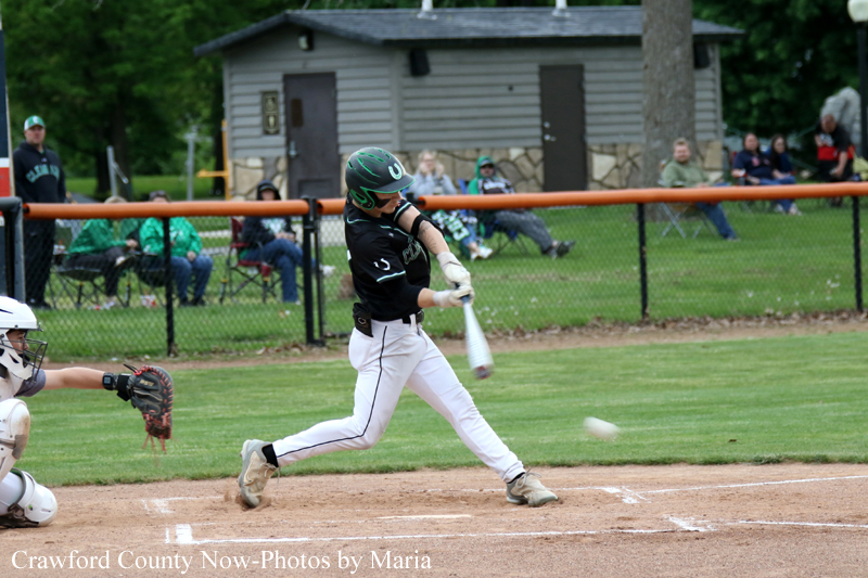 Young baseball batter in a black jersey swings at a pitch on a dirt infield as the ball approaches.