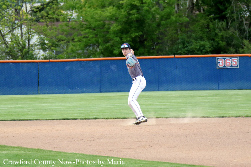 Baseball player in gray jersey and white pants stands in a fielding position on the dirt infield, glove ready, with a blue outfield wall behind him and a '365' sign in the background.