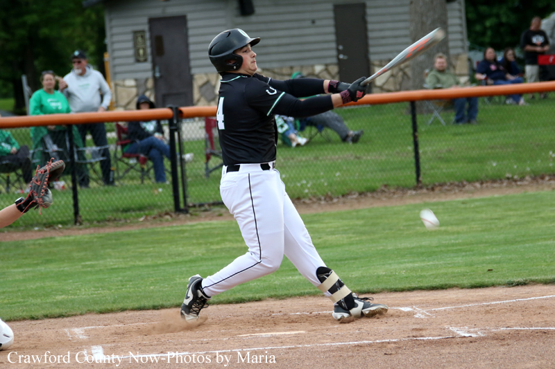 Female softball batter in a black jersey and white pants swings at a pitched ball during a game, with a catcher’s glove visible to the left and spectators in the background.