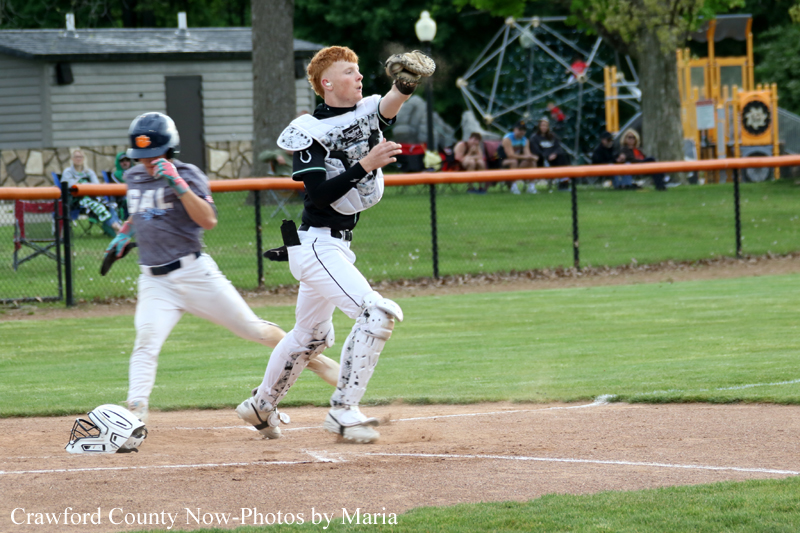 Baseball catcher in white gear extends glove toward home plate as a runner slides in, crowd watching in the background.