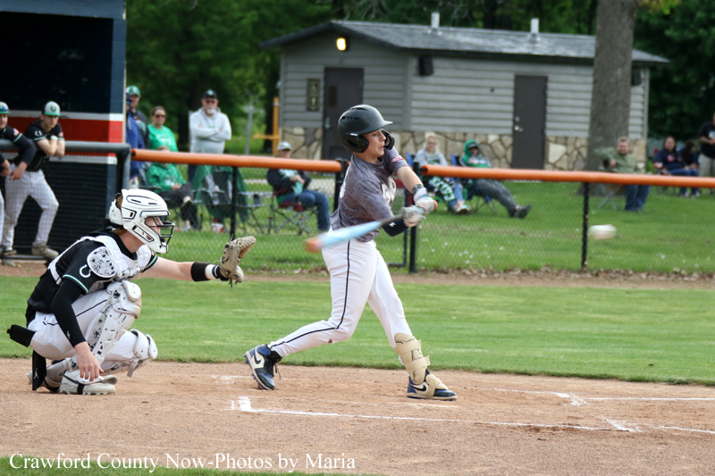 Baseball action at home plate: batter swings in gray uniform as the catcher crouches behind the plate and the ball is in midair.