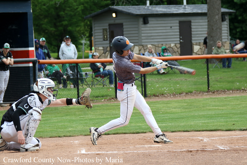 Baseball batter in a gray shirt swings at a pitch, with the catcher in black and white crouched behind him on the dirt infield.