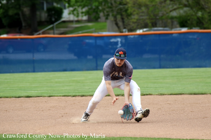 Baseball player in gray shirt and white pants lunges to field a ground ball on the infield, glove reaching toward the ball with a blue fence in the background.