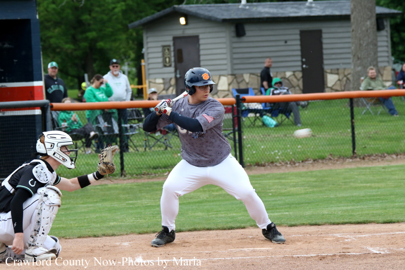 Baseball batter in gray shirt and white pants swings at a pitch while catcher crouches behind home plate on a dirt infield.