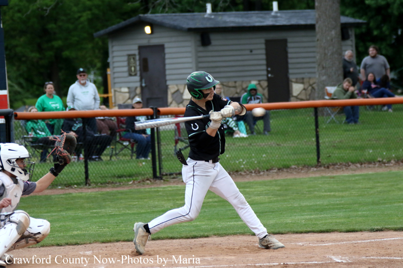 Baseball batter in a black uniform swings at a pitch, with a catcher in white gear behind him and spectators in the background.