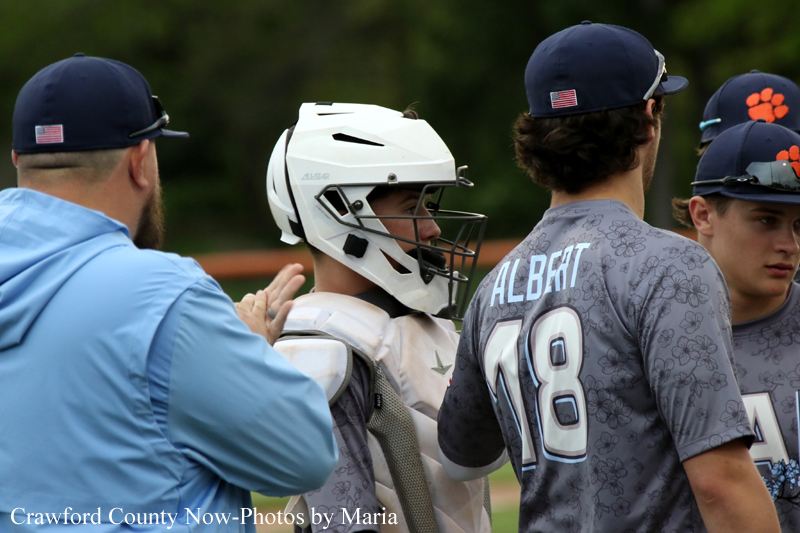 Baseball players chat on a field: a catcher in white helmet and chest guard stands with a teammate wearing a gray jersey reading 'Albert' and number 18; coach in blue reaches in.