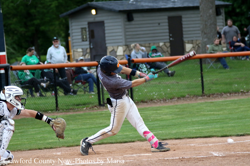 Baseball batter in gray uniform swinging at a pitch, catcher ready behind him, with spectators in the outfield."