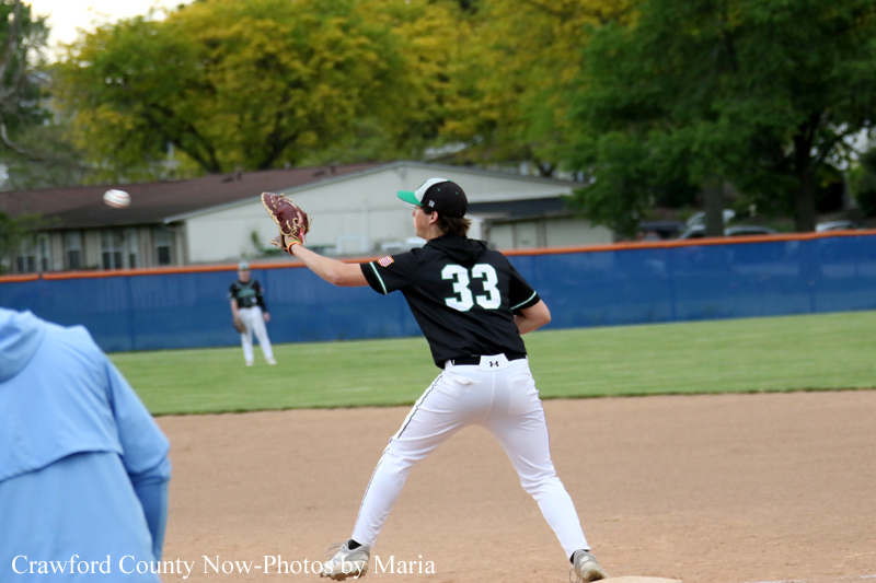 Baseball pitcher in black uniform #33 winds up to throw from the mound on a sunny field with a blue outfield fence behind him.