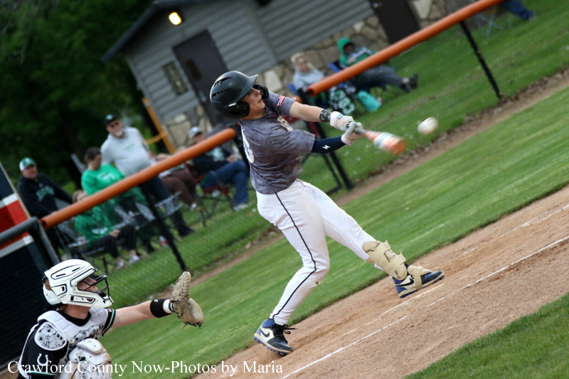 Baseball batter in a gray jersey swings at a pitch while the catcher watches from home plate; spectators sit beyond the fence.
