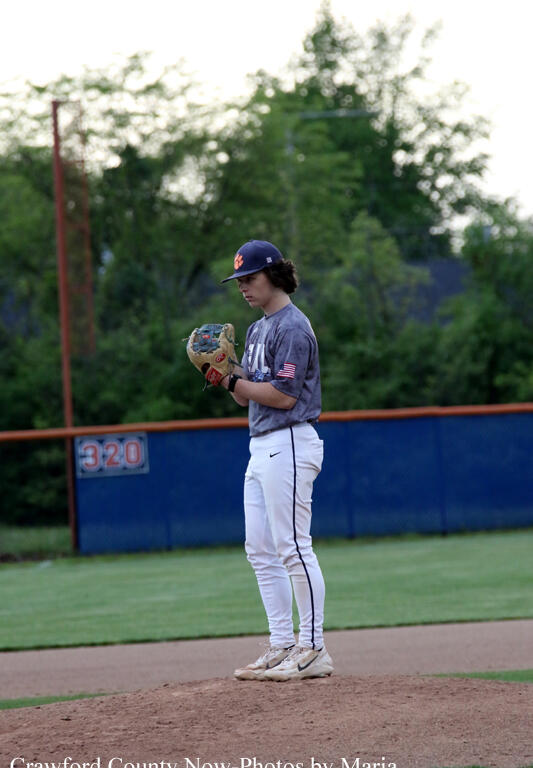 Young baseball pitcher stands on the mound wearing a gray jersey, white pants, and a navy cap, glove ready for a pitch.