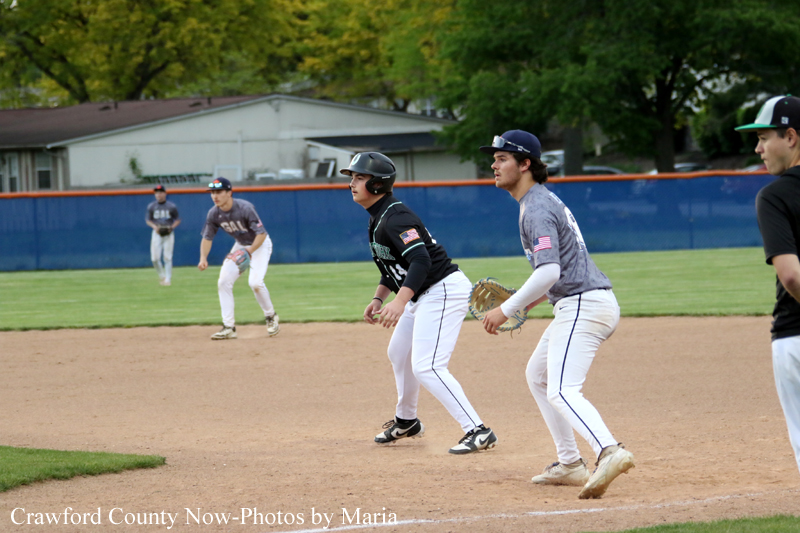 Baseball infield action: players positioned and ready to field on a dirt diamond with a blue fence in the background.