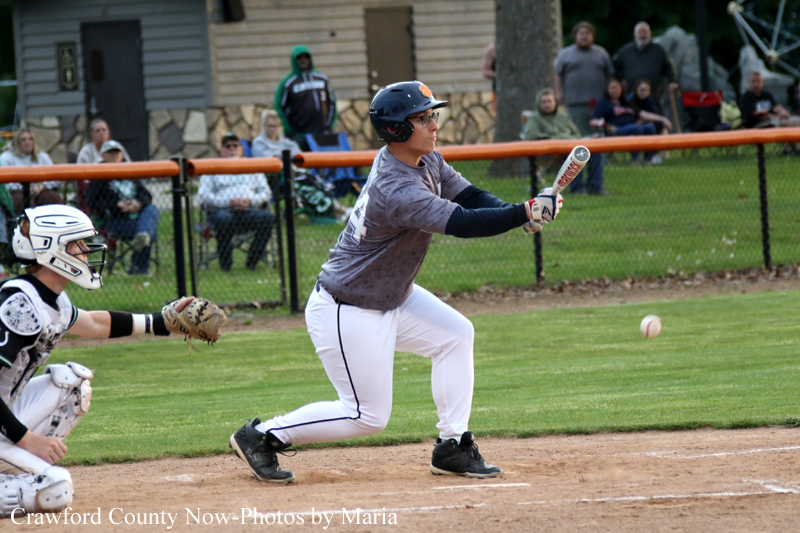 Baseball batter in a helmet and gray shirt swings at a pitch during a game, with the catcher and spectators in the background.