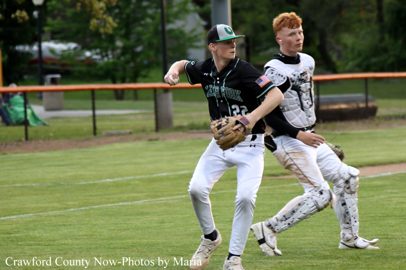 Two baseball players are in action on a grassy field: one in a dark uniform throwing a glove-side pitch, the other wearing catcher gear chasing behind him.