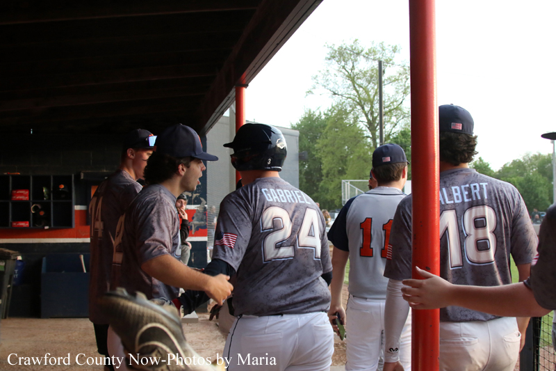 Baseball players in gray uniforms stand near the dugout under a pavilion, backs to the camera.