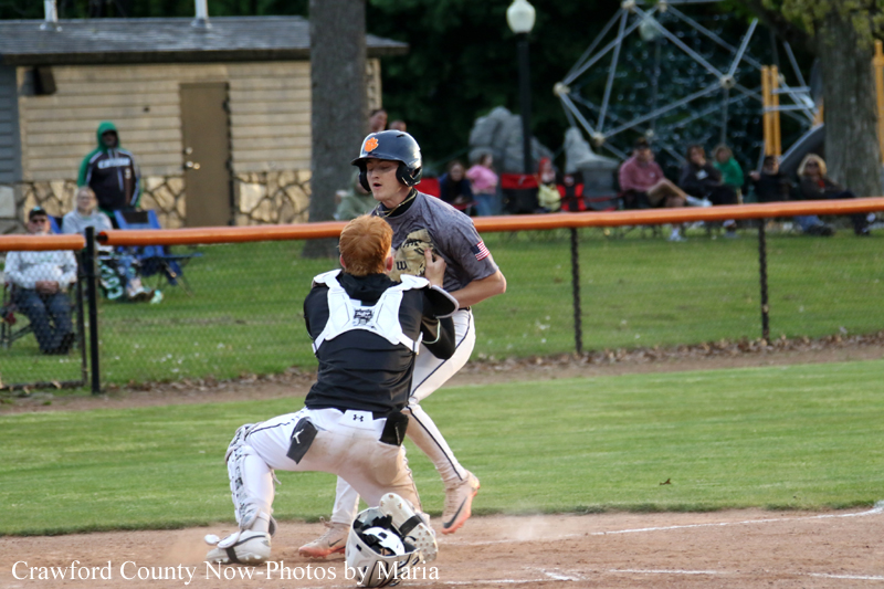 Baseball catcher in white gear crouches behind home plate as a baserunner in gray jersey advances.