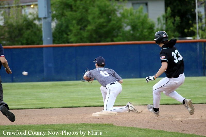 Baseball infield action: a runner slides into a base as a fielder reaches to tag, with another player running nearby on the field behind.