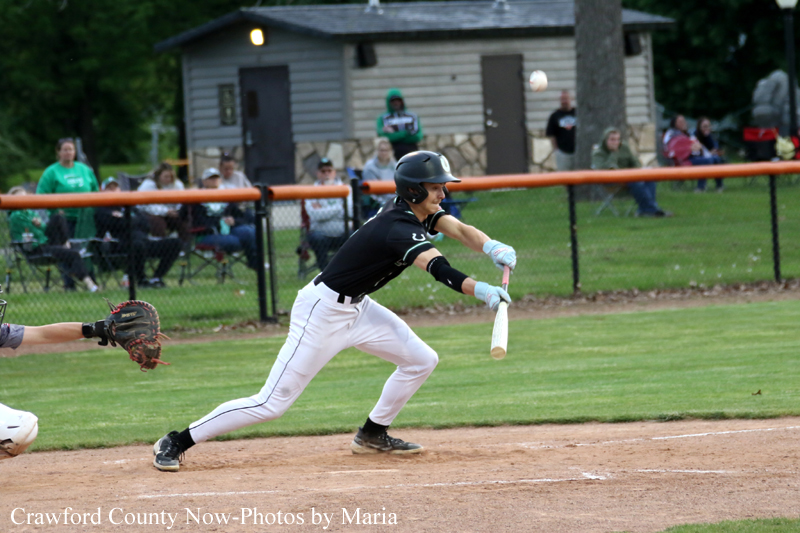 Baseball batter in black uniform swings at a pitched ball while a catcher reaches from left, on a dirt infield with a grassy outfield and spectators in the background.