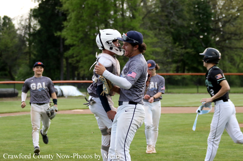 Baseball coach hugs a catcher on a grassy field as teammates celebrate in the background.